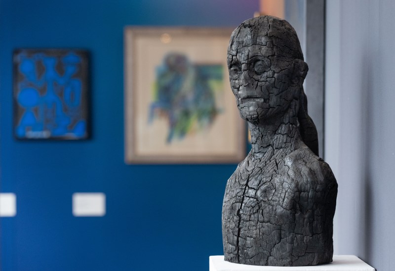 Statue einer zerbrochenen Frau in Grau, im Hintergrund zwei Gemälde