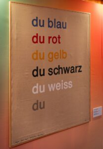 Bild mit den Farben Blau, Rot, Gelb, Schwarz und Weiß, in deutscher Sprache geschrieben und in den entsprechenden Farben gemalt.