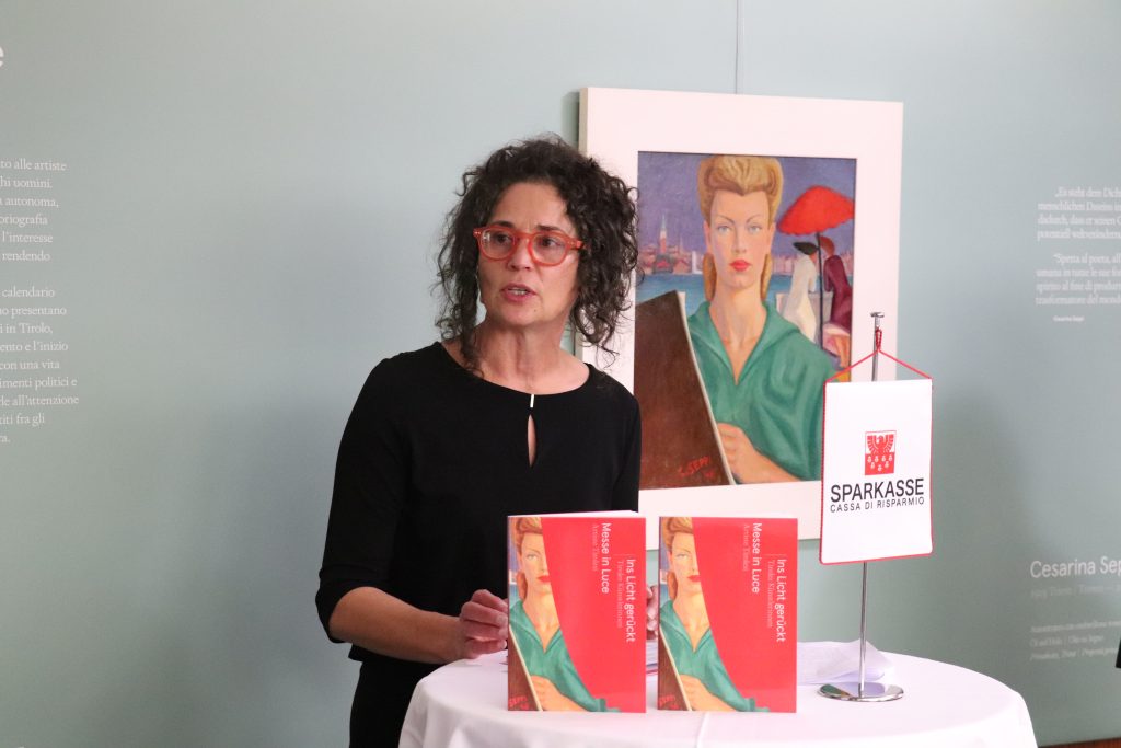 Silvia Höller mit zwei roten Büchern vor sich und einem Gemälde mit der Darstellung einer Frau hinter sich